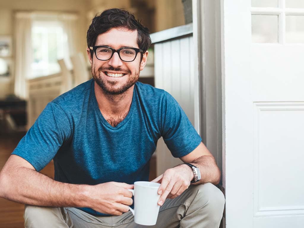 Un bărbat cu ochelari și o cămașă albastră stă pe podea, ținând o ceașcă de cafea în mâini