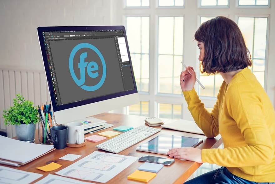 O femeie stă la birou, concentrată pe ecranul calculatorului, cu un caiet și un pix alături