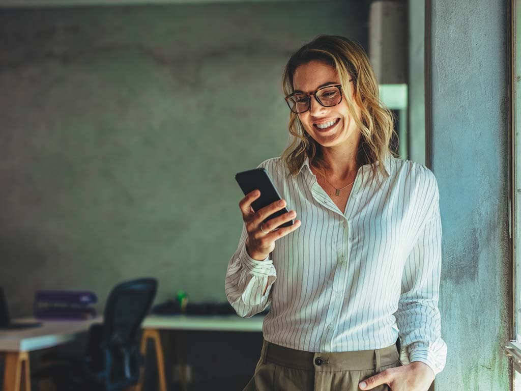 O femeie cu ochelari ține un telefon mobil, uitându-se la ecran cu o expresie concentrată