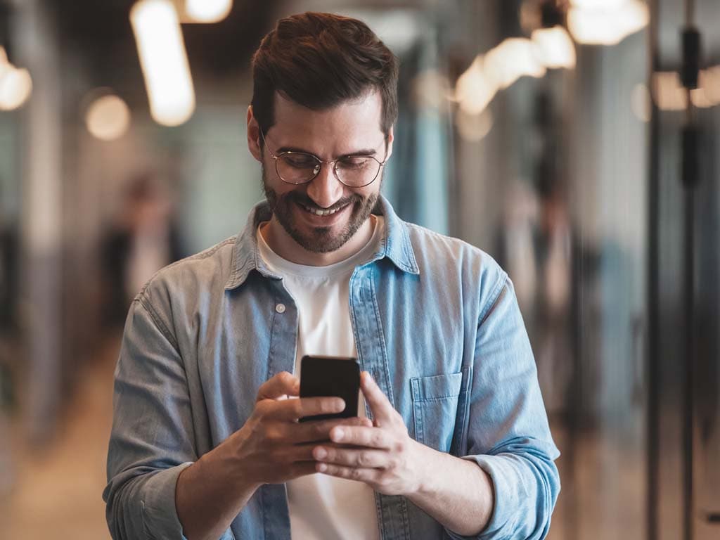 Un bărbat cu o cămașă albastră zâmbește în timp ce privește telefonul, părând implicat și fericit