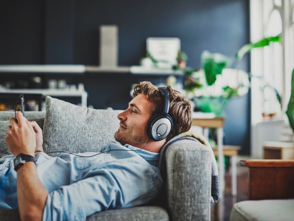 Un bărbat stă pe o canapea purtând căști, concentrat asupra telefonului în timp ce se relaxează