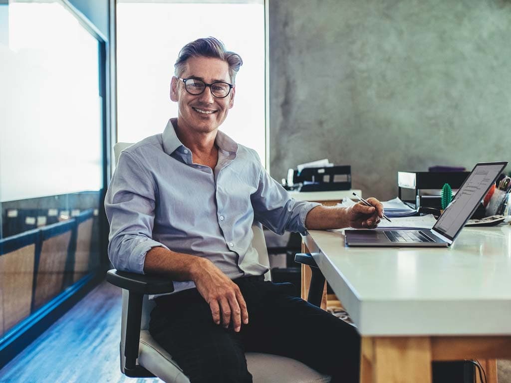 Un bărbat zâmbitor, așezat la birou, lucrând la un laptop, emanând o atitudine pozitivă și implicată