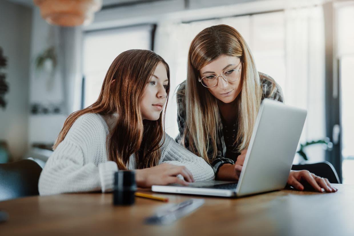 Două femei concentrate asupra unui laptop, discutând informațiile afișate pe ecran