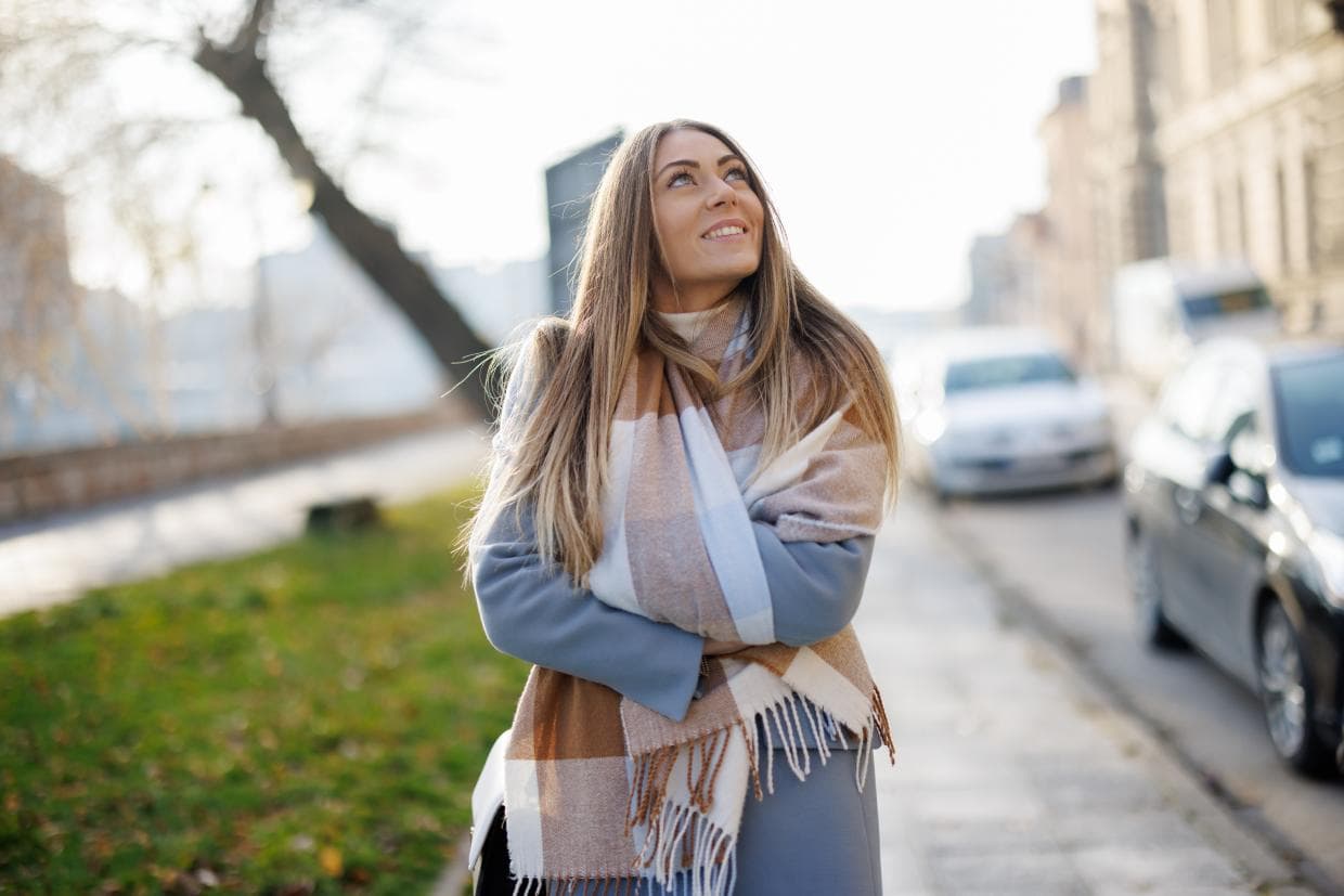 O femeie în palton și eșarfă stă pe stradă, înconjurată de peisaj urban, părând gânditoare și calmă