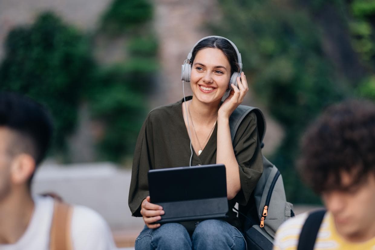 O femeie care poartă căști folosește un laptop în timp ce stă pe o bancă într-un spațiu public