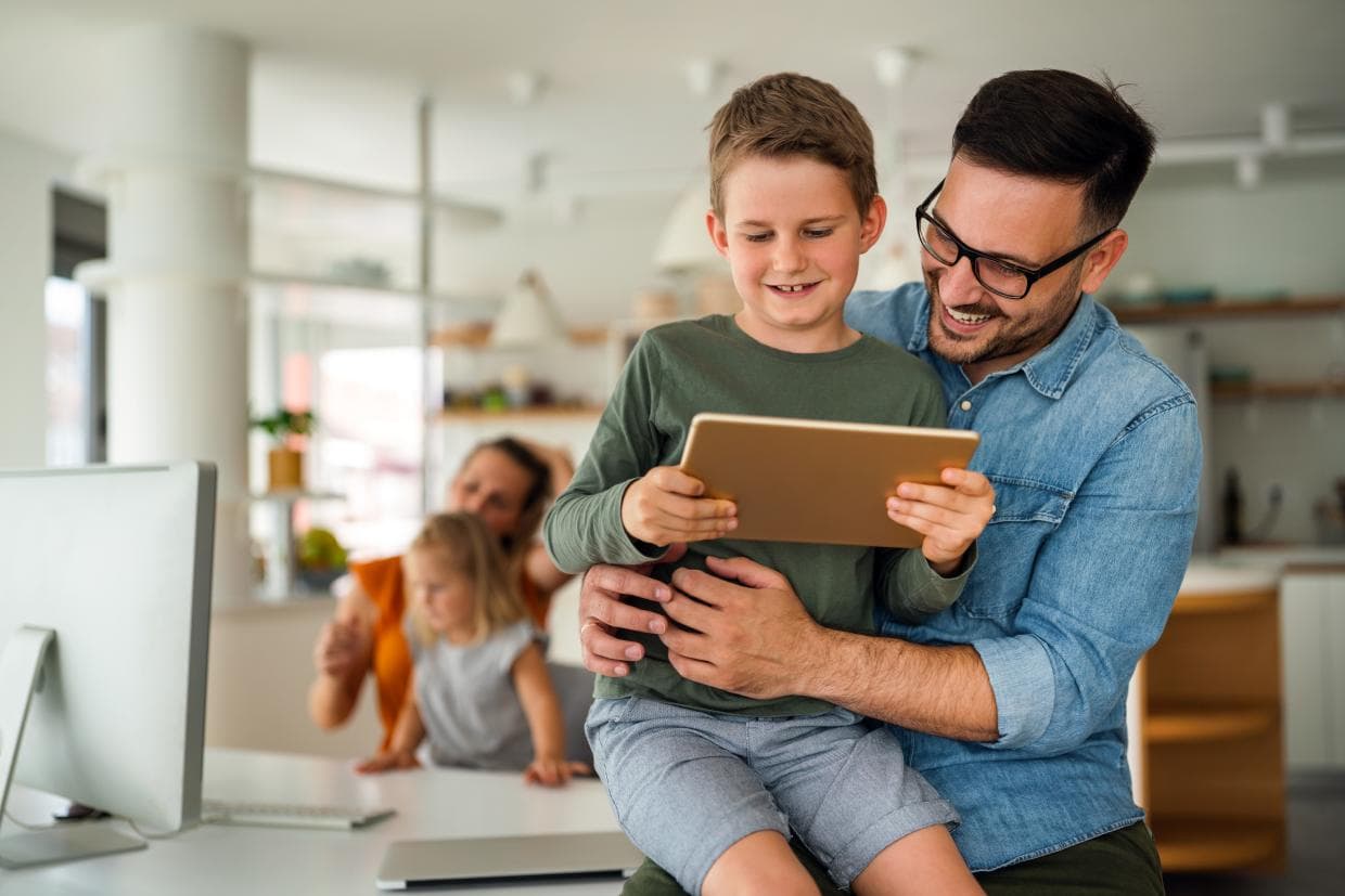 Un bărbat și un băiat stau împreună pe canapea, concentrați asupra unei tablete din mâinile lor