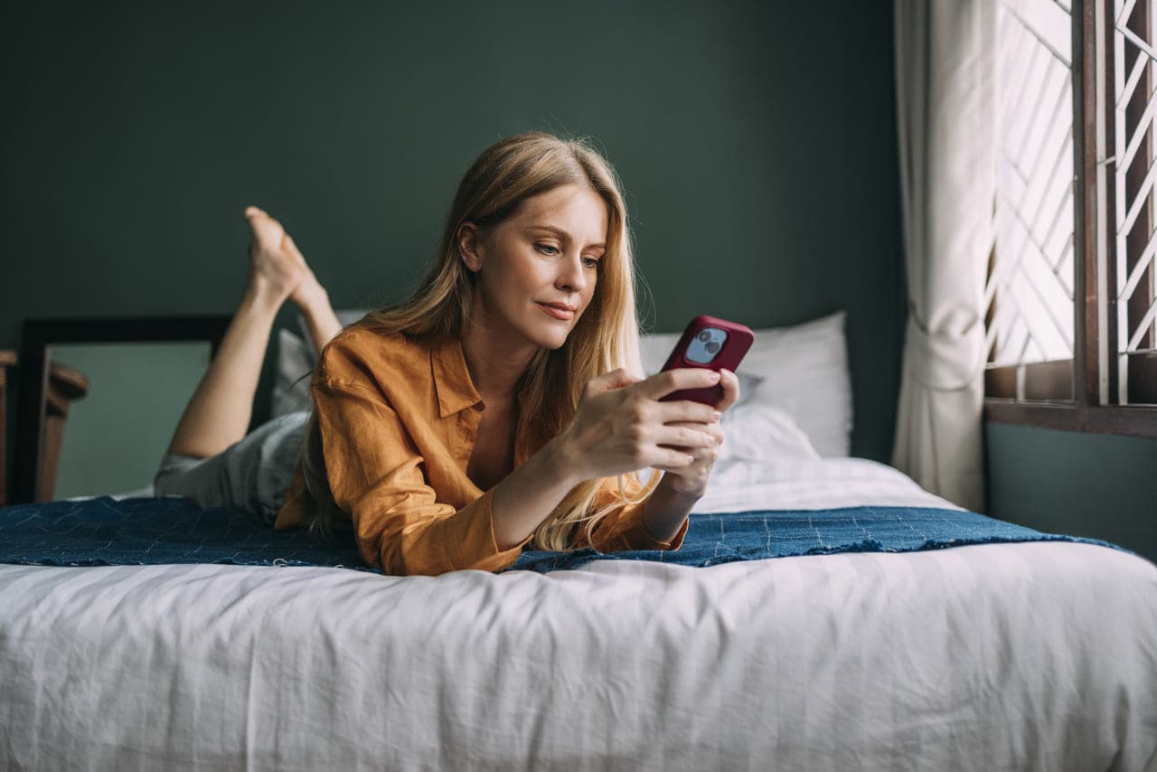 O femeie stă întinsă pe pat, concentrată pe telefon, cu o expresie relaxată și așternuturi moi în jurul ei
