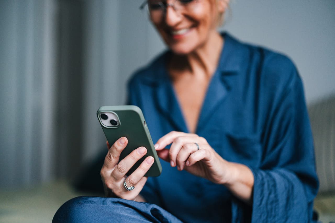 O femeie în pijamale stă și își folosește telefonul; pare relaxată și interesată de dispozitiv