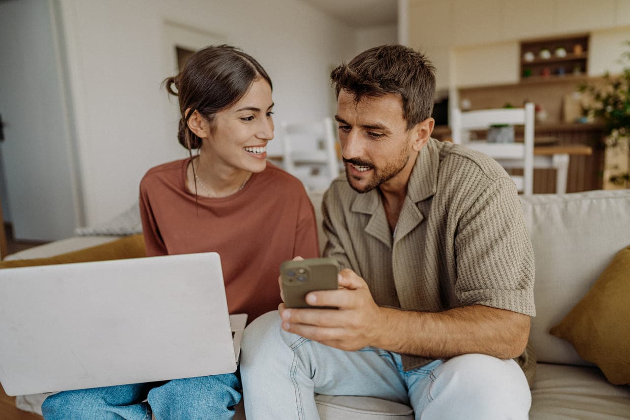 Un cuplu stând pe o canapea, folosind împreună un laptop și un smartphone într-un mediu casnic confortabil
